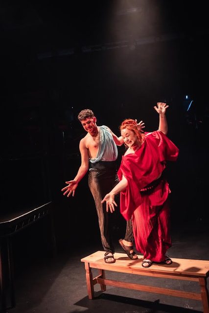 Jess in a red robe in a dramatic two-hander scene under stage lighting