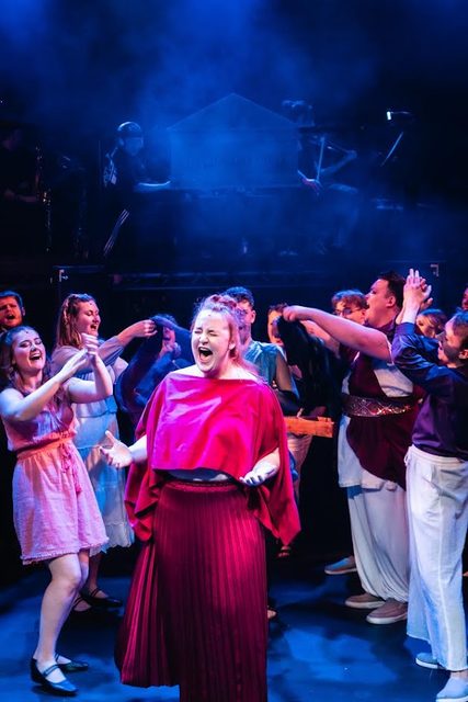 Jess singing centre stage in a magenta costume with an ensemble behind her